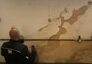 Ein Mann untersucht eine feuchte Wand mit abgeplatztem Putz und freiliegenden Rohren.